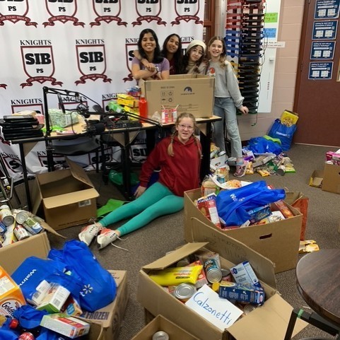 This photo features Vinu P., Ayat, Lana, Harper, and Everleigh in a room with boxes of food donations. 